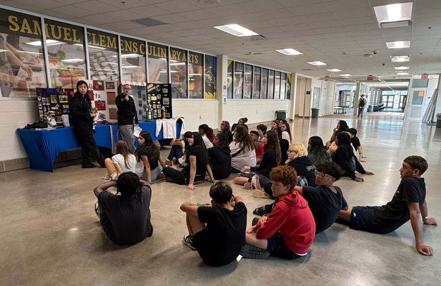 junior high students sit in a high school hallway, observing a presentation by culinary arts. a paneled glass window in back reads "samuel clemens culinary arts”
