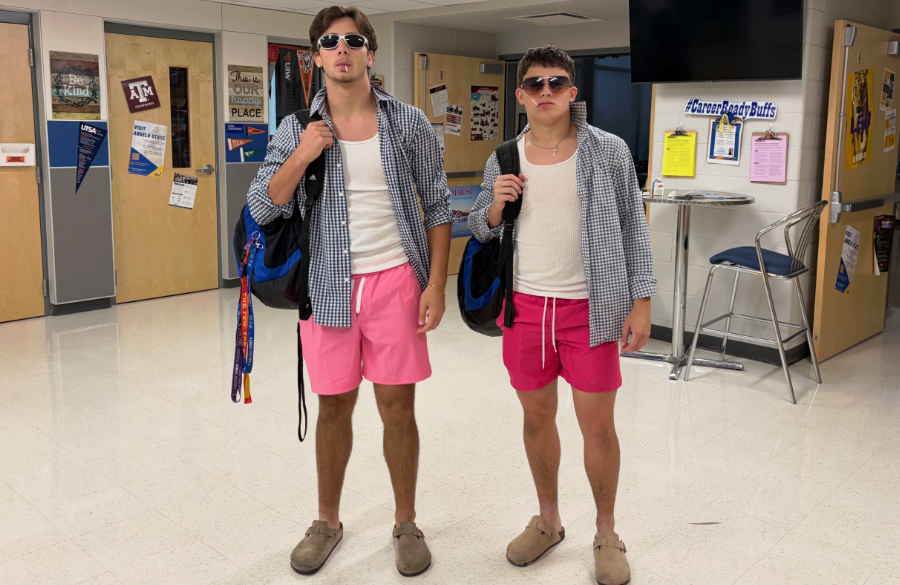 two male high school students are dressed identically in matching beige shoes, no socks, pink shorts, white t-shirts, and a long-sleeved checkered button-up shirt. each student has a blue backpack slung over the right shoulder, wears sunglasses, and has a lollipop in their mouths.