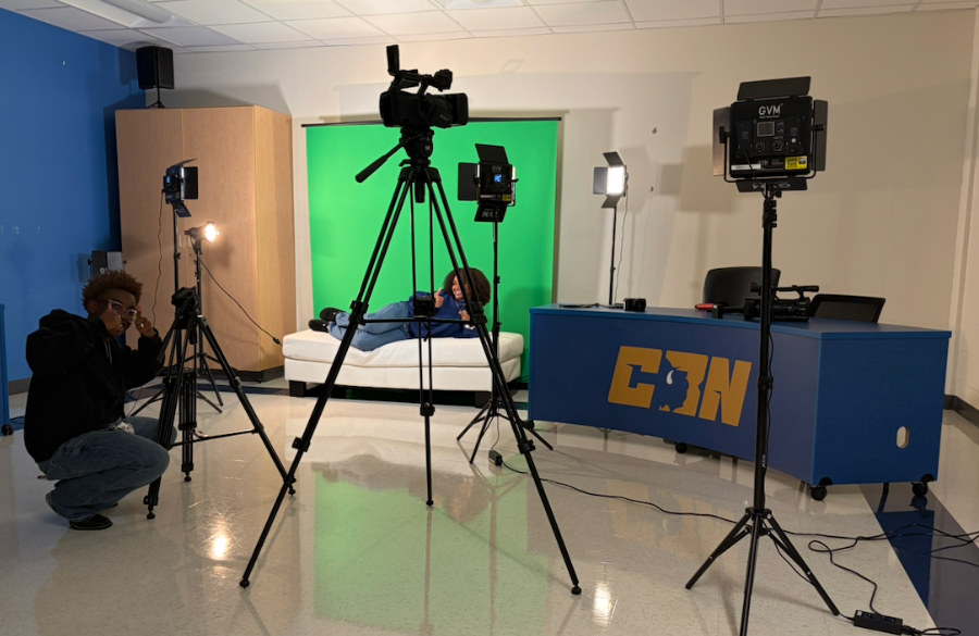 high school classroom converted into a broadcast studio. present in the darkened room are several lights and a video camera on tripods. one student at left with fingers pointed to his head while another student reclines on a white sofa in background, positioned in front of a green screen. at right is a blue news desk with the letters "cbn" centered in a yellow font.
