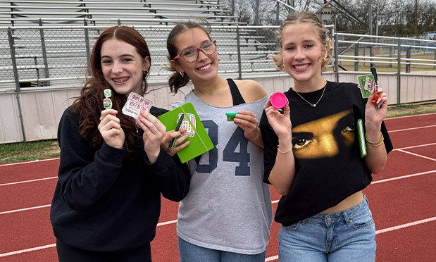 three high school girls smile while standing on a running track, each holding trinkets promoting mental health