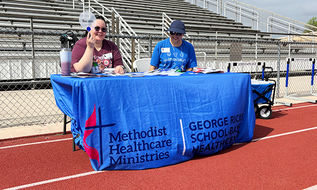 two women sit at a table adorned with a blue cover. on it is the methodist cross & flame symbol, along with text "methodist healthcare ministries: george ricks school-based healthcare." one of the women holds up a fan.