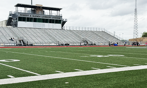 wide-angle shot of a high school football field, track, and bleachers beneath a cloudy sky. students walk on the track in the background. signage on the press box at the top of the bleachers reads "robert & glenda lehnhoff stadium"