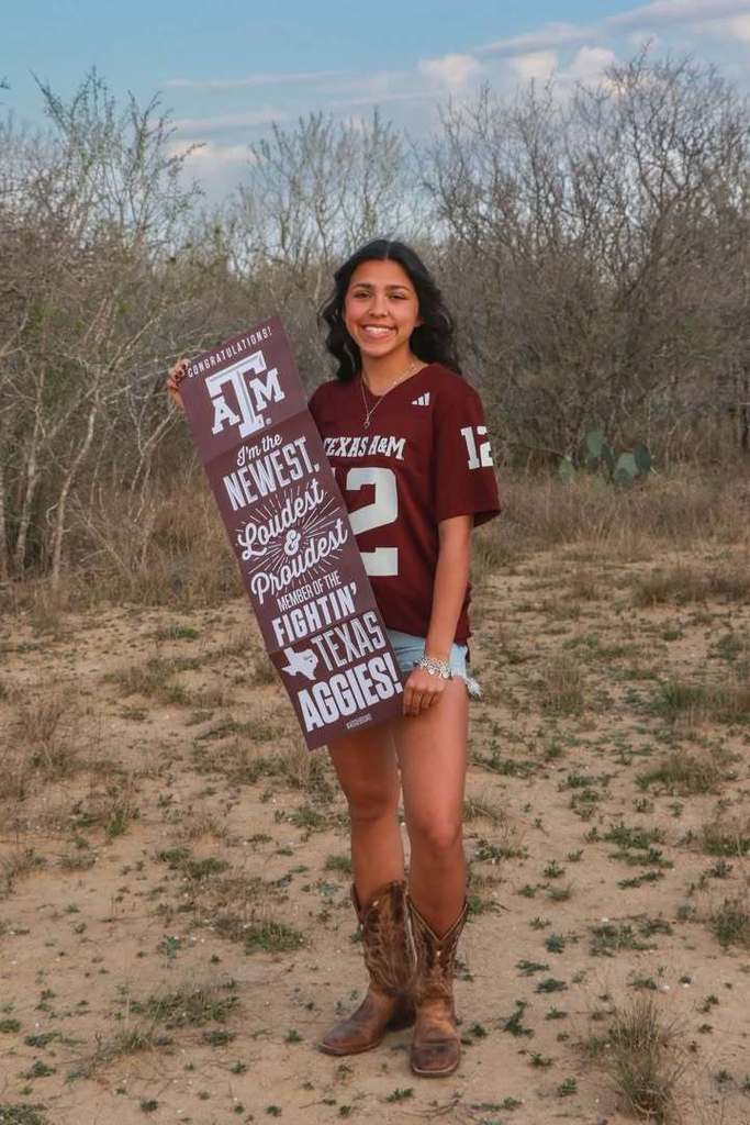 a high school student stands in a rustic clearing wearing cowboy boots, jean shorts, and a texas a&m university maroon jersey with the number 12 on it. the student holds a sign with the texas a&m university logo with text reading "i'm the newest, loudest, and proudest member of the fightin' texas aggies!"