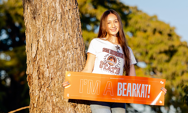 a high school student wearing a sam houston state t-shirt leans against a tree beneath a cloudless sky holding a sign which reads "i'm a bearkat!"