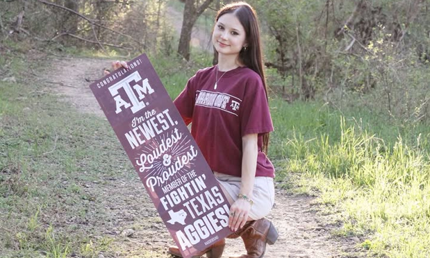 a high school student kneels along a wooded pathway, holding a banner with the texas a&m university masthead and text reading "i'm the newest, loudest, & proudest member of the fightin' texas aggies!" the student wears cowboy boots, a kahki skirt, and a maroon texas a&m university shirt