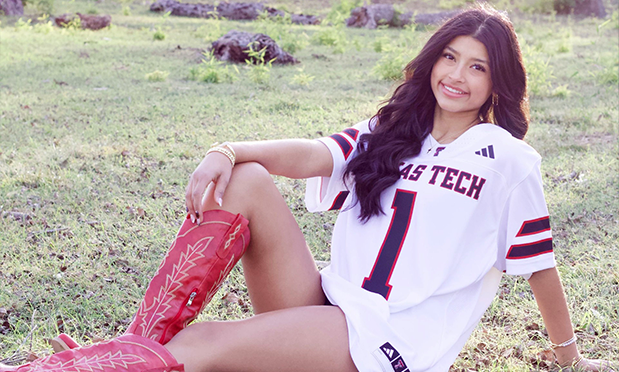 a high school senior sits in a grassy field wearing red cowboy boots and a white texas tech jersey with the number "1" on it