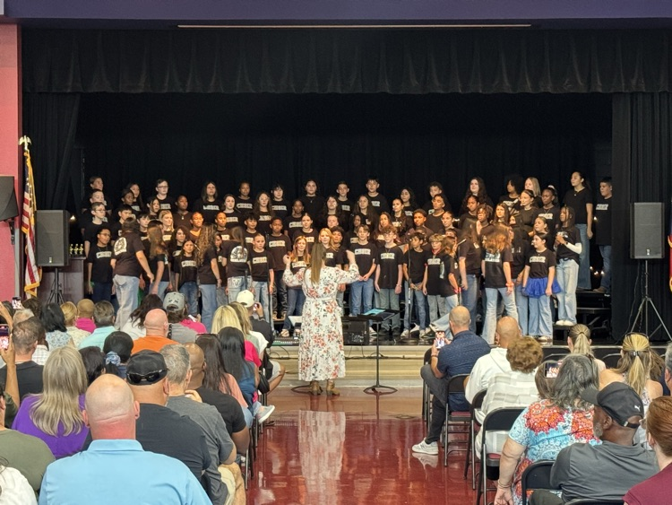 in the cafeteria, the crowd watches the entire choir upon the stage. Well, Miss Alvarado is conducting.