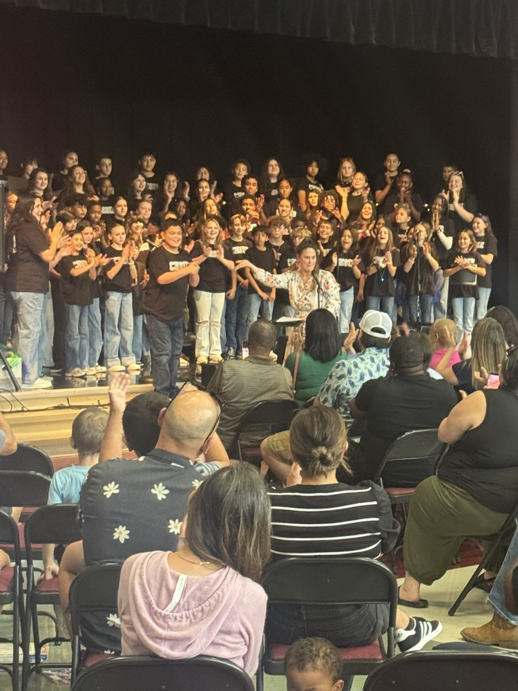 The choir is clapping as a student is standing next to Miss Alvarado and she is having her hand out in a presentation towards the boy