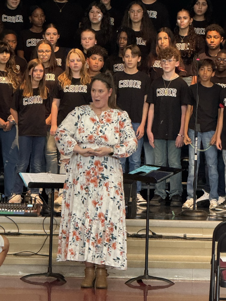Miss Alvarado is standing in front of the choir up on the stage and she is making a  singing face