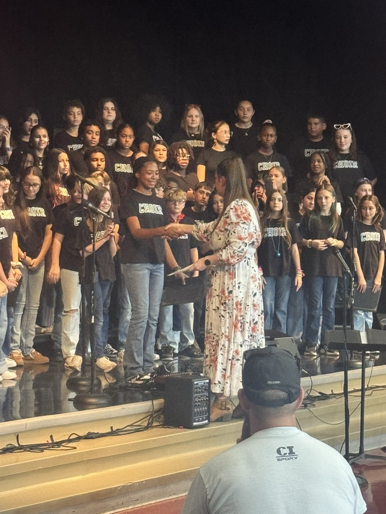 Alvarado shakes a student’s hand, giving her an award with the choir standing behind