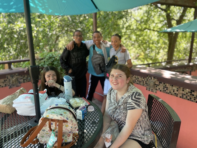 Two girls sitting at a table with three boys standing behind them at lunch in the shade