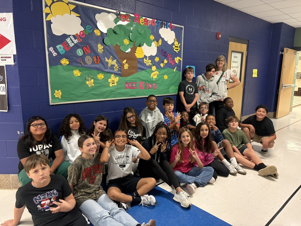 Mrs. Rindahl and her class sitting in the hallway beneath the bulletin they decorated with a tree landscape and bees, saying " I beelieve in you. bee kind. bee responsible, 