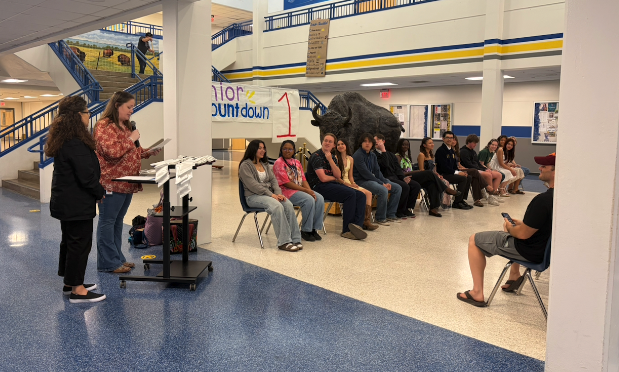 a high school teacher stands at a podium, speaking into a microphone while students and their parents listen
