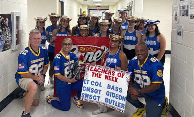 high school dance team in sparkling hats and uniforms flank three kneeling adults in football jerseys. a held sign reads "teachers of the week: lt col bass, cmsgt gideon, smsgt butler"