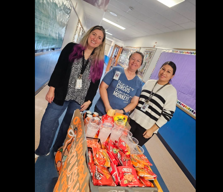 Three teachers in the blue hallway standing near the cart with orange snacks