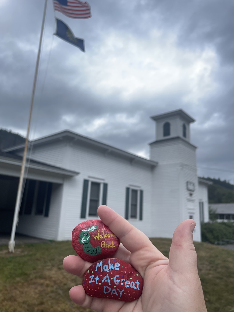 Hand holding painted rock in front of school.  Rock says Welcome Back and Make it a Great Day