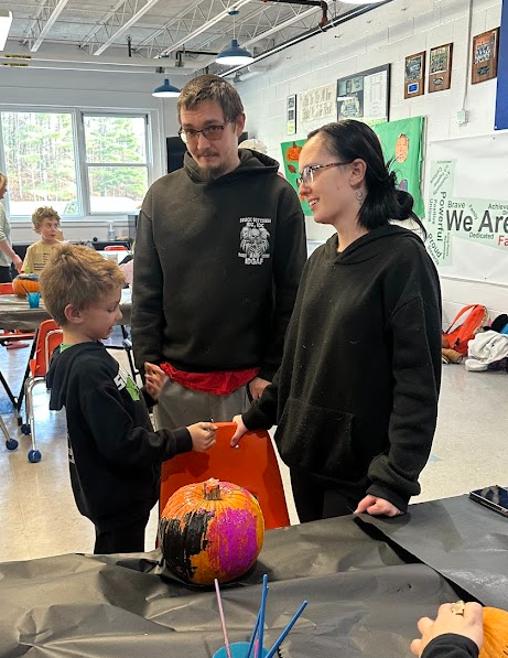 A young student proudly shows a brightly painted pumpkin to two adults who are smiling and chatting with him. They are standing beside a table covered in art supplies inside a bright school room with large windows and colorful posters on the wall.