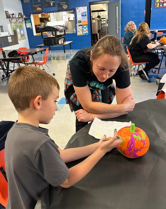 A student works carefully on a colorful pumpkin while an adult leans over with a smile, watching and encouraging him. They are seated at a table covered with a black cloth in a bright school cafeteria, with more families and pumpkins in the background.