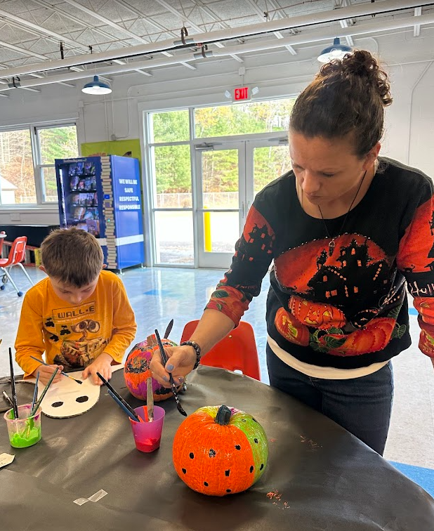 A student and an adult paint pumpkins together at a table in a bright school space. The adult is wearing a colorful Halloween sweater with pumpkins on it, and the student is focused on adding details to his creation. Paintbrushes and small paint cups sit nearby, and large windows show trees outside.