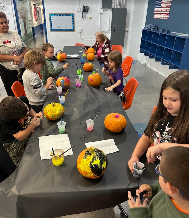 A group of young students sit around a long table covered with a black tablecloth, painting small pumpkins with bright colors. Paint cups, brushes, and napkins are scattered across the table. Two adults stand nearby supervising with smiles. The room appears to be a school hallway with orange chairs and cubbies in the background.