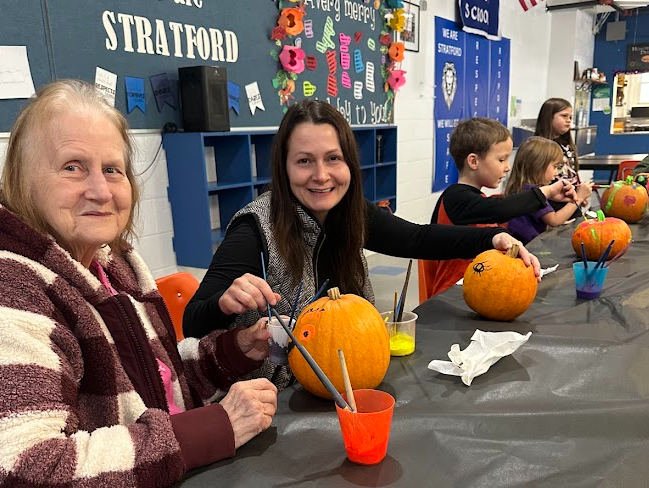 Two adults sit side-by-side at a table, smiling as they paint pumpkins. Colorful cups of paint and brushes are spread out in front of them. Several students are also visible down the table, focused on decorating their own pumpkins. A bulletin board and school banners hang on the wall behind them.