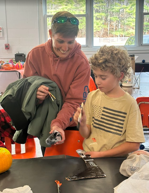 A smiling adult and a student paint together at a table, holding paintbrushes and working with black paint. A small pumpkin sits nearby on the black table covering. Sunlight comes in through the large windows behind them, and finished pumpkins can be seen on a table in the background.