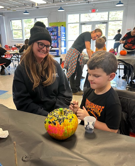 AA smiling adult wearing a winter hat sits beside a young student as he paints a brightly colored pumpkin. They are seated at a table covered with a black cloth in a sunny school space, with other families painting pumpkins in the background.