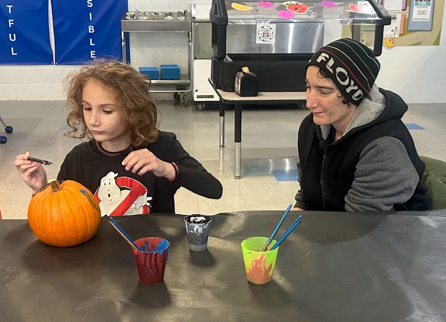 A student with curly hair carefully paints a pumpkin while an adult sits beside them watching proudly. Several cups of paint and brushes sit on the table covered in a black tablecloth. The scene takes place in a school cafeteria with posters on the wall in the background.
