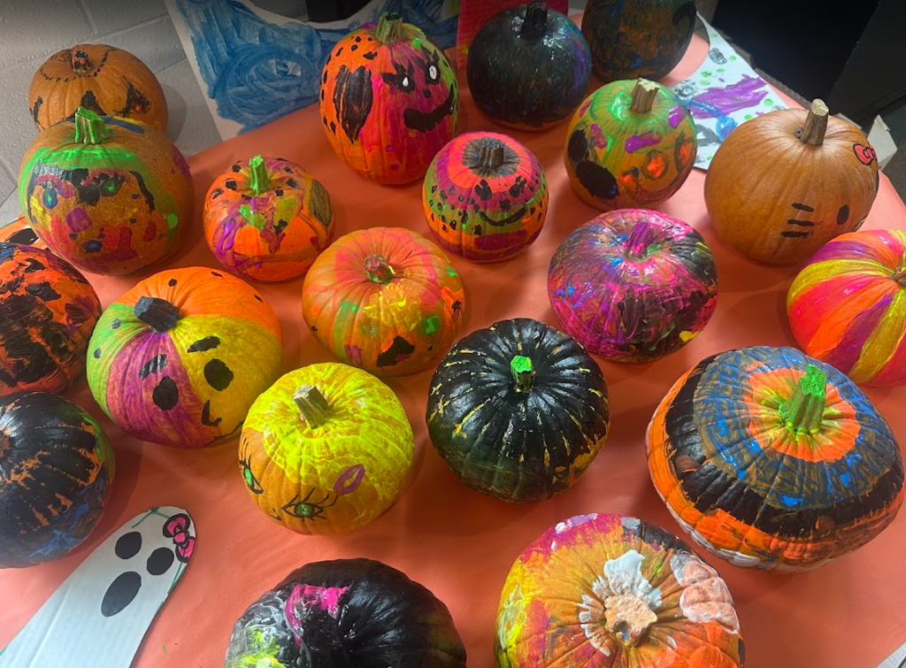 A table covered with an orange tablecloth displaying a collection of small pumpkins painted by students. The pumpkins are decorated with bright neon colors, bold black paint, expressive faces, polka dots, stripes, and creative patterns. A small paper ghost drawing with a bow is placed near the front. The background shows student artwork taped to the wall.