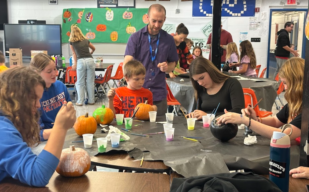 A large group of students sit at several tables painting pumpkins with colorful paints. A staff member stands nearby offering help. Cups of paint, brushes, and pumpkins in various stages of decoration fill the tables. More families are visible in the background, and painted pumpkin artwork is displayed on a green bulletin board behind them.
