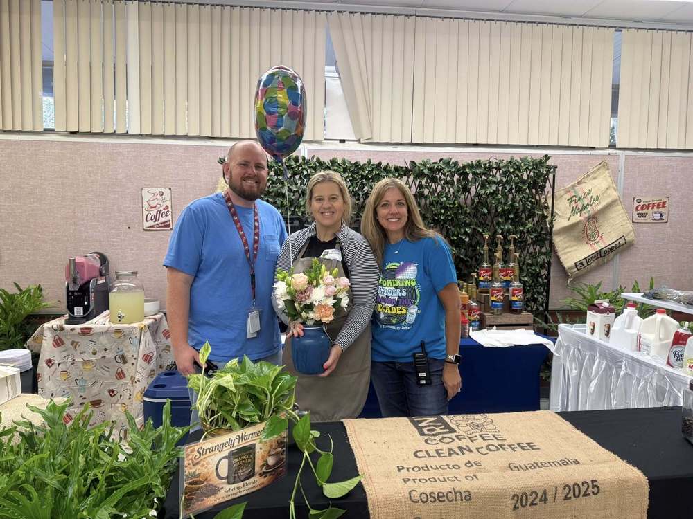  Gemini said Three people stand smiling behind a coffee display table decorated with burlap sacks and greenery. The woman in the center holds a bouquet of flowers and a balloon. The setup includes coffee syrups, creamers, and a sign for "Strangely Warmed" coffee in Sebring, Florida.