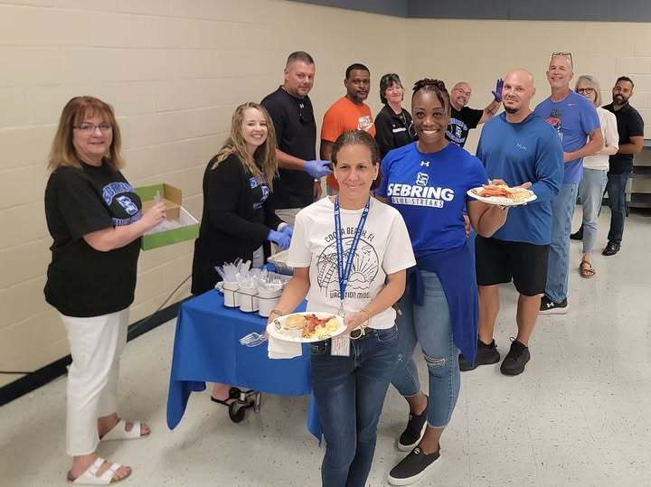 Sebring High School staff smile while serving and receiving food at a welcome-back event. A long line of teachers and staff stretches across the room, holding plates of food. A table with utensils and condiments sits at the front, creating a friendly and festive atmosphere.