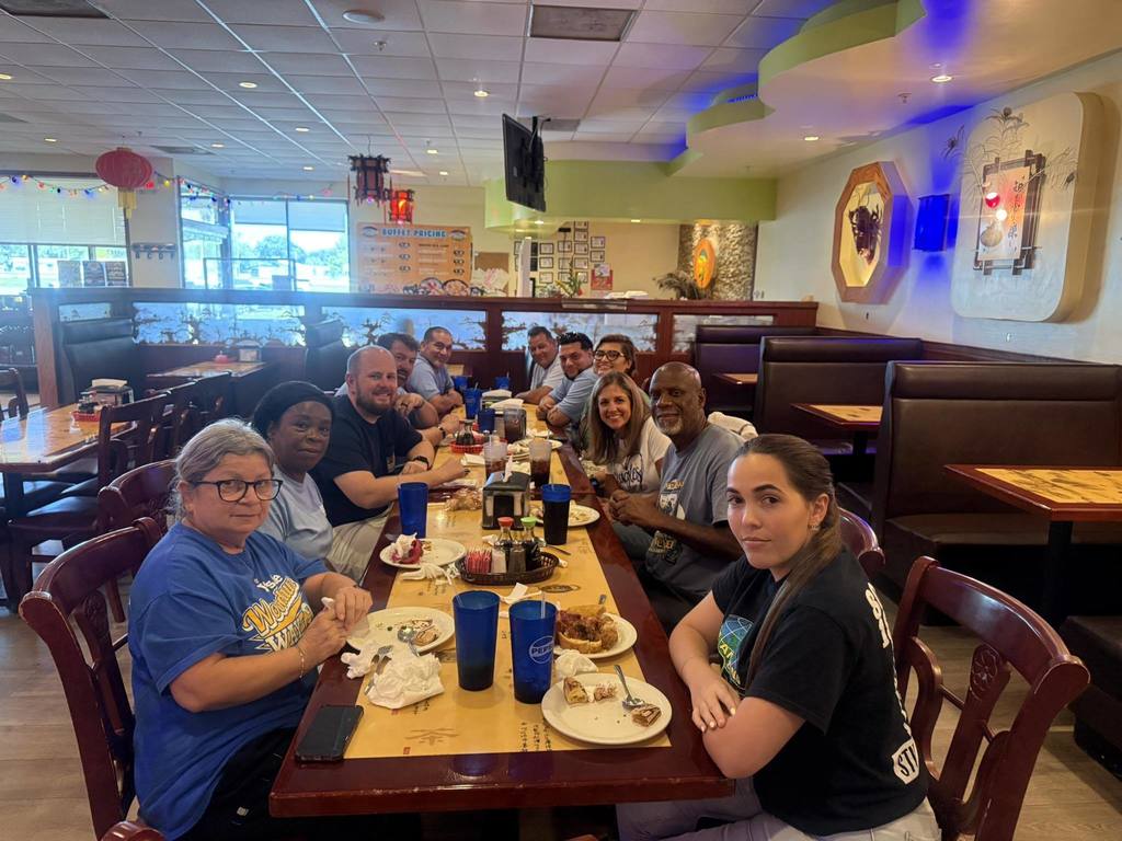  School staff sit together around a long restaurant table, enjoying a meal and smiling for the camera. Plates of food and drinks are in front of them, and the atmosphere is casual and cheerful.