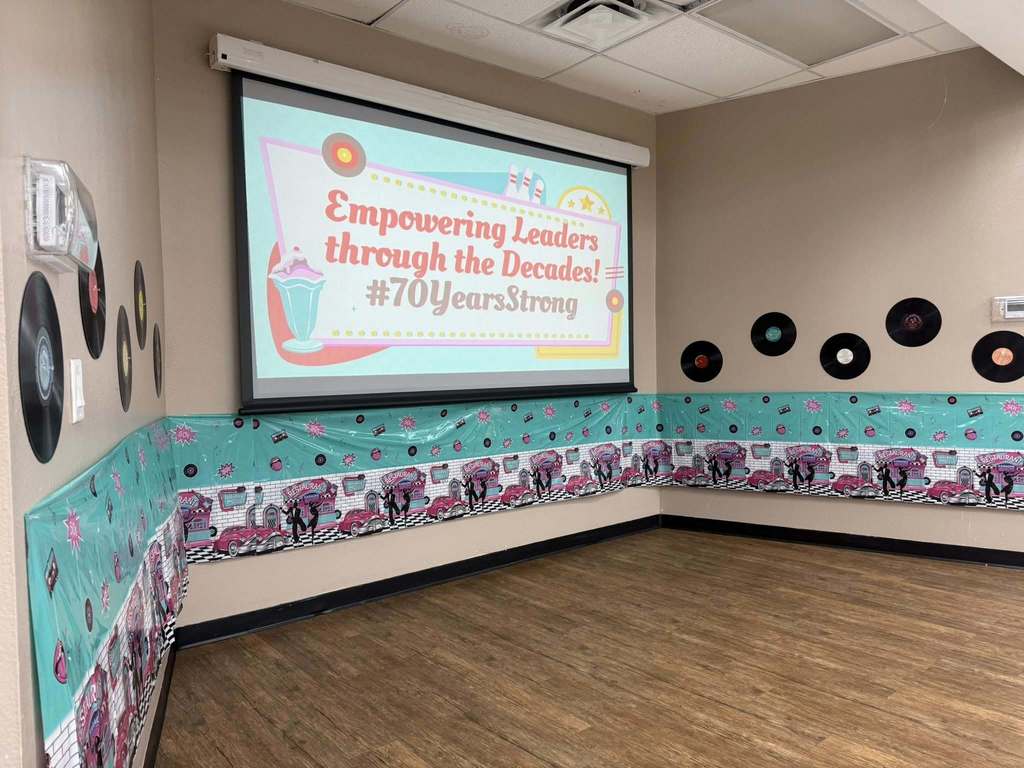A decorated room with a retro 1950s theme features a large screen that reads “Empowering Leaders through the Decades! #70YearsStrong.” The walls are adorned with colorful diner-style decorations, silhouettes of dancers, and vinyl records, creating a festive and nostalgic atmosphere.