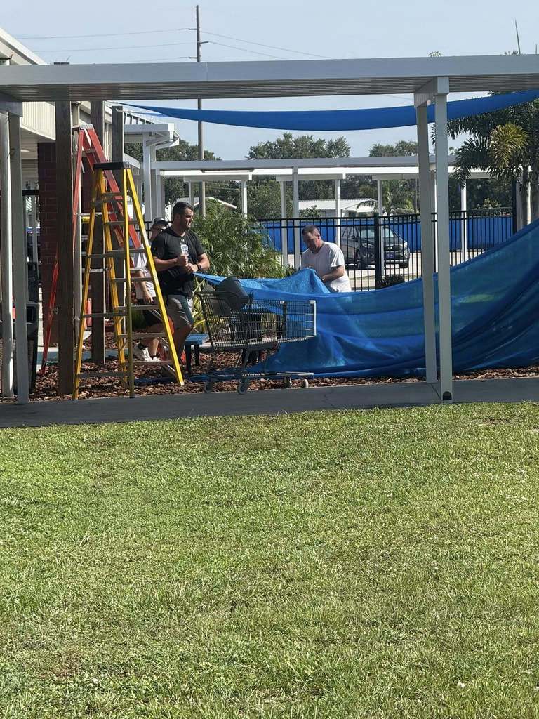  Two men work to secure a large blue tarp to a metal frame structure, with one man standing on a ladder and the other on the ground. A shopping cart sits nearby, holding the excess tarp. The scene appears to be an outdoor construction or maintenance project on a sunny day.