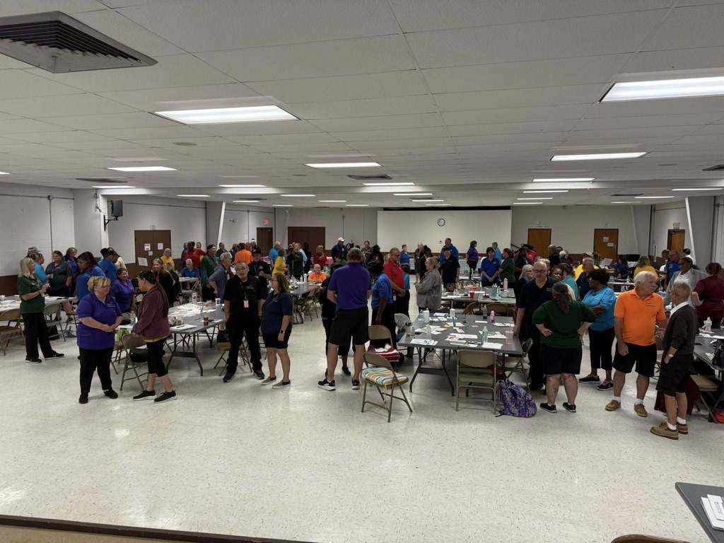 A large group of people gather in a spacious multipurpose room with tables and chairs. Many are standing and talking in small groups, wearing colorful shirts. The environment appears to be a workshop, training, or team-building event with materials spread across tables.