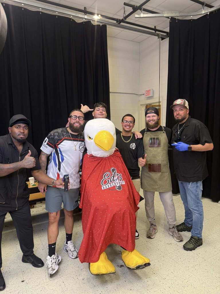 Six men pose with a person in an eagle mascot costume wearing a red barber cape labeled "Ed's Barbershop." Some of the men hold barber tools, and they are smiling in a room with black curtains and white walls, suggesting a fun event or community haircut day.