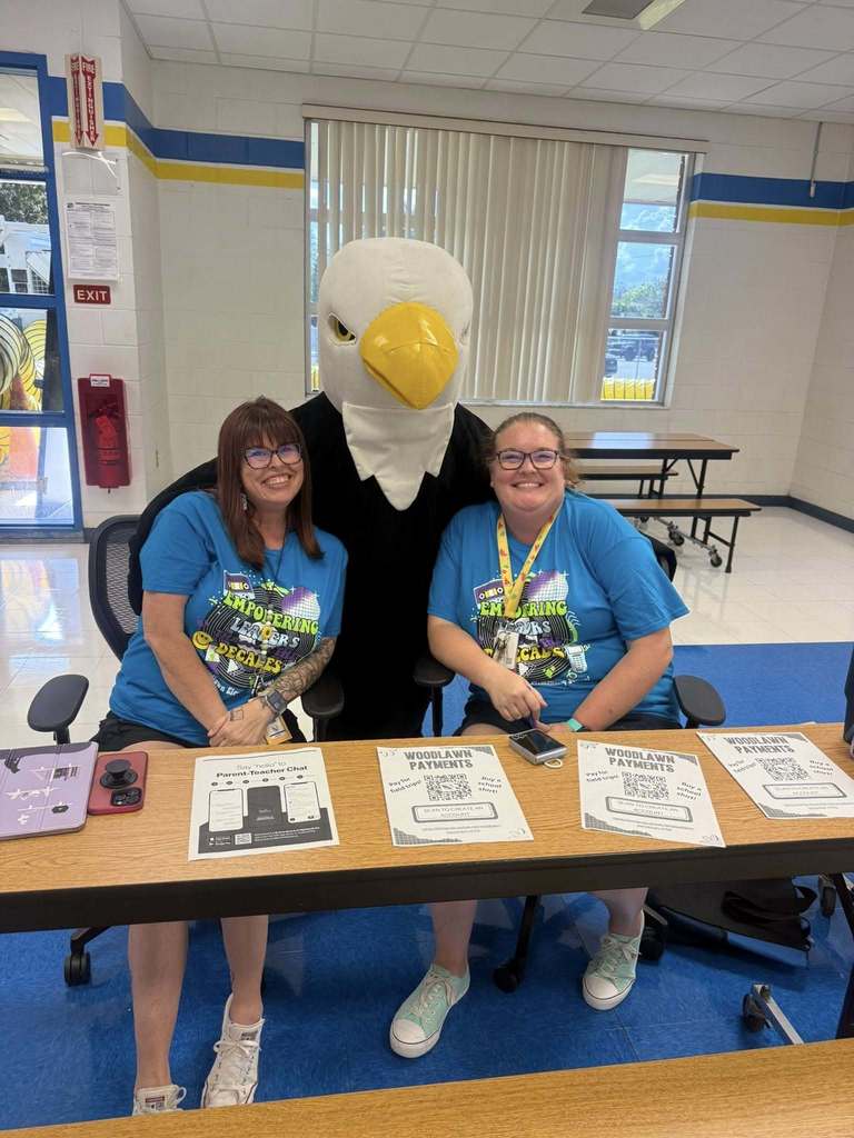 Two smiling women in matching blue shirts sit at a table with flyers and QR codes, flanked by a person in an eagle mascot costume. The setting is a bright, cheerful school room with blue and yellow accents and large windows in the background.