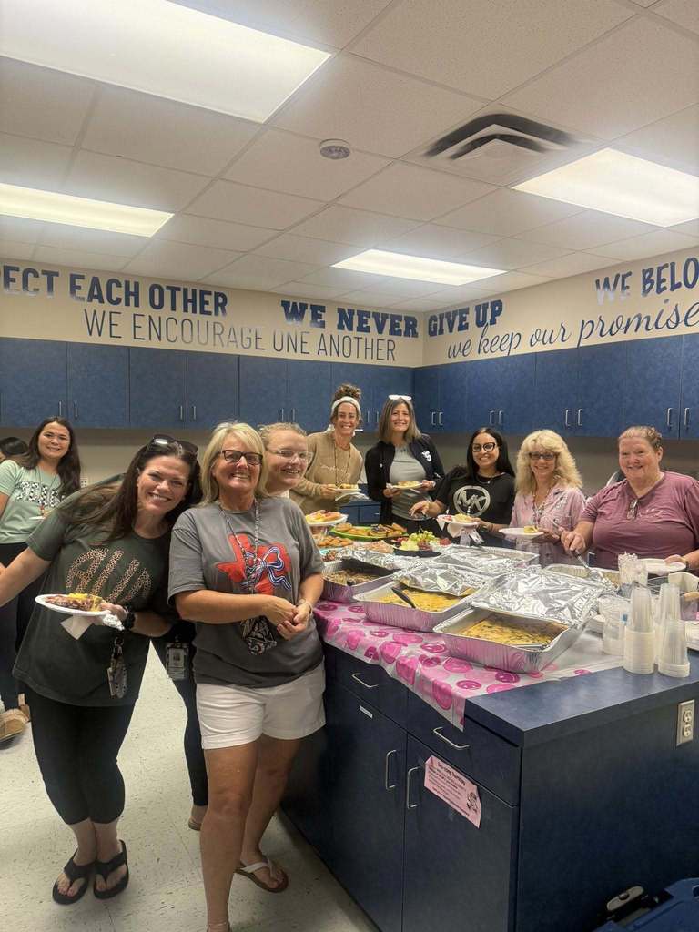  A group of smiling women, likely teachers, stand behind a long table filled with a potluck meal. They are gathered in a classroom with motivational posters on the wall that read "RESPECT EACH OTHER," "WE ENCOURAGE ONE ANOTHER," "WE NEVER GIVE UP," and "WE KEEP OUR PROMISES."