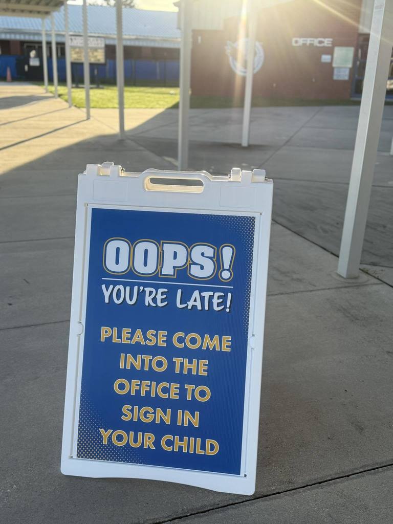 A sandwich board sign outside a school reads, “OOPS! YOU’RE LATE! Please come into the office to sign in your child” in white and yellow text on a blue background. Sunlight shines from behind, with the school office building visible in the background.