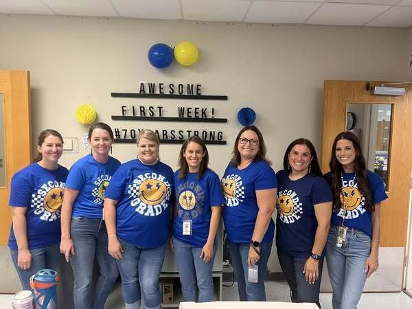Seven smiling teachers wearing matching blue “Second Grade” shirts stand together in a classroom. Behind them, a wall display reads “AWESOME FIRST WEEK! #70YearsStrong” with blue and yellow decorations.