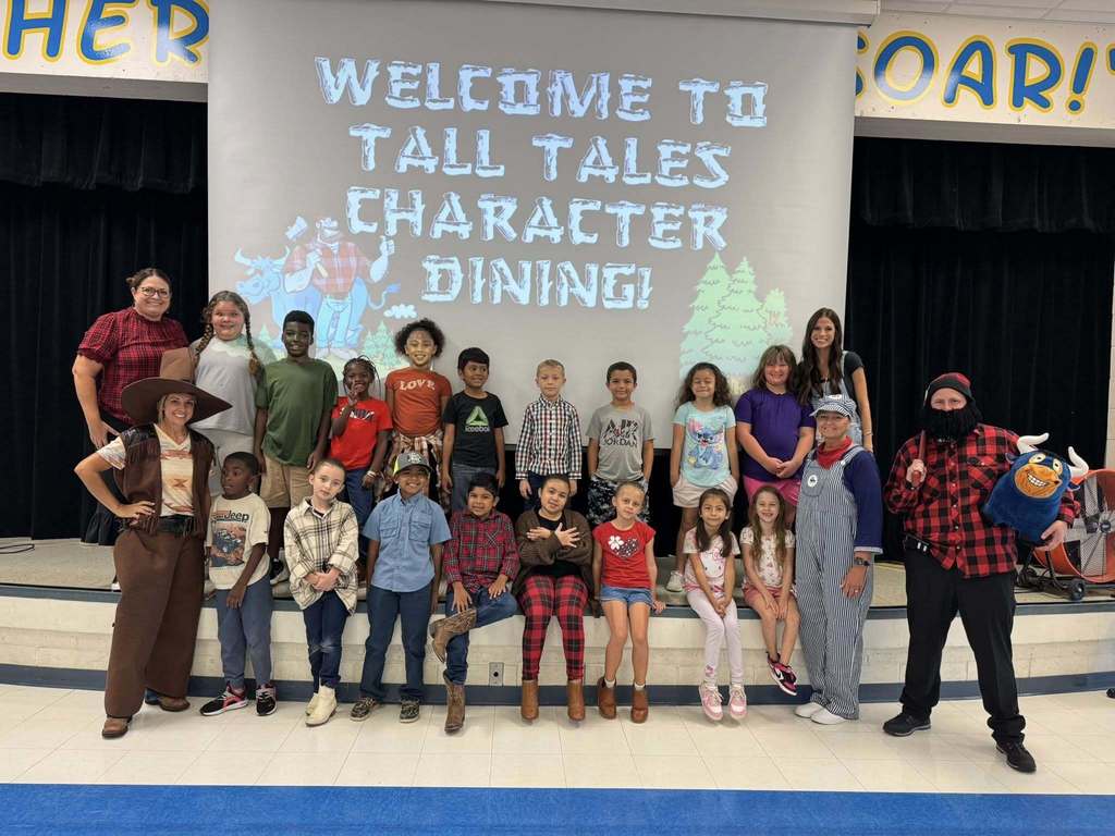 A group of elementary students and teachers pose on stage in front of a screen reading “Welcome to Tall Tales Character Dining!” Some adults are dressed as tall tale characters, including a cowboy, lumberjack, and train conductor. Students wear casual clothes and smile together for the photo.