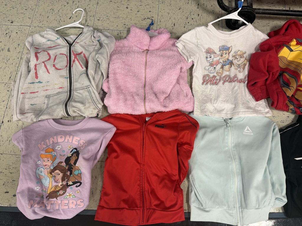 A collection of lost and found items from a school. Numerous children's jackets and hoodies are displayed on a clothing rack and on the floor. The collection also includes a variety of water bottles, lunch boxes, a hair bonnet, noise-canceling earmuffs, and jewelry laid out for identification.