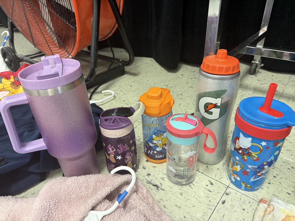 A collection of lost and found items from a school. Numerous children's jackets and hoodies are displayed on a clothing rack and on the floor. The collection also includes a variety of water bottles, lunch boxes, a hair bonnet, noise-canceling earmuffs, and jewelry laid out for identification.