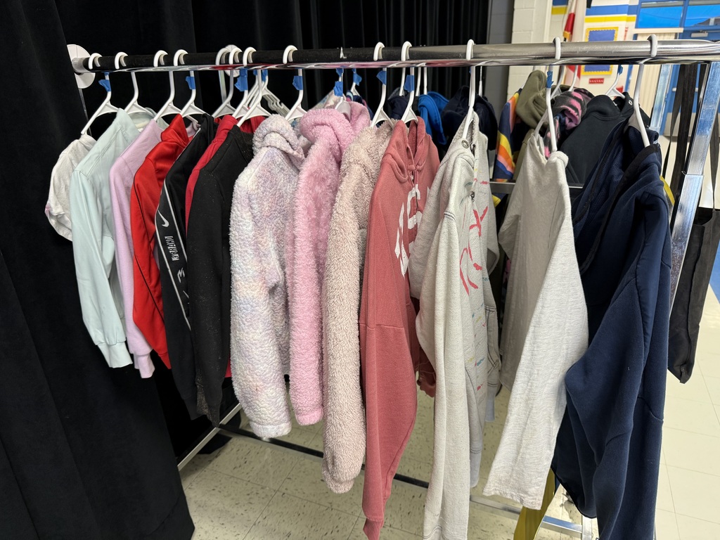 A collection of lost and found items from a school. Numerous children's jackets and hoodies are displayed on a clothing rack and on the floor. The collection also includes a variety of water bottles, lunch boxes, a hair bonnet, noise-canceling earmuffs, and jewelry laid out for identification.