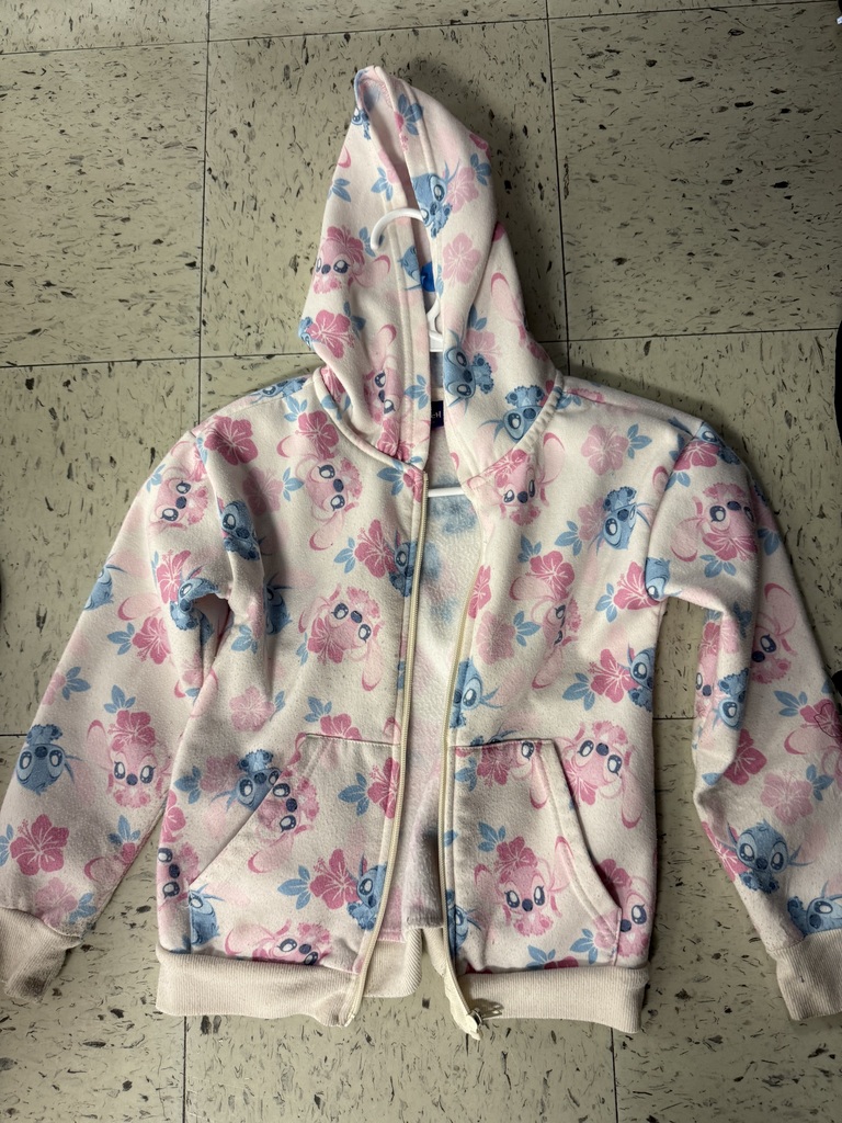 A collection of lost and found items from a school. Numerous children's jackets and hoodies are displayed on a clothing rack and on the floor. The collection also includes a variety of water bottles, lunch boxes, a hair bonnet, noise-canceling earmuffs, and jewelry laid out for identification.