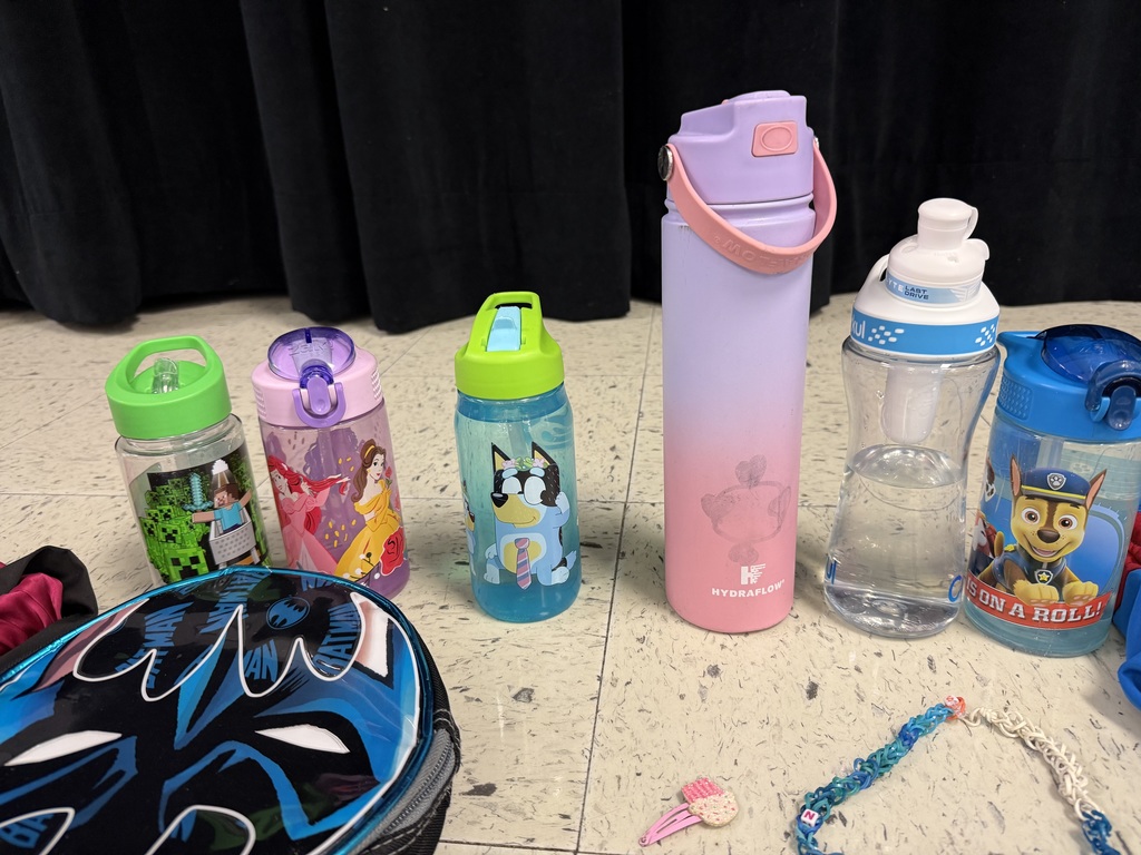 A collection of lost and found items from a school. Numerous children's jackets and hoodies are displayed on a clothing rack and on the floor. The collection also includes a variety of water bottles, lunch boxes, a hair bonnet, noise-canceling earmuffs, and jewelry laid out for identification.
