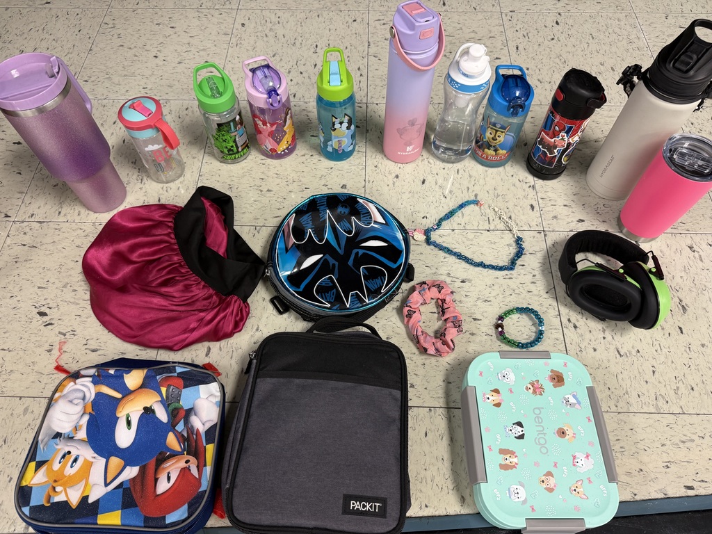 A collection of lost and found items from a school. Numerous children's jackets and hoodies are displayed on a clothing rack and on the floor. The collection also includes a variety of water bottles, lunch boxes, a hair bonnet, noise-canceling earmuffs, and jewelry laid out for identification.