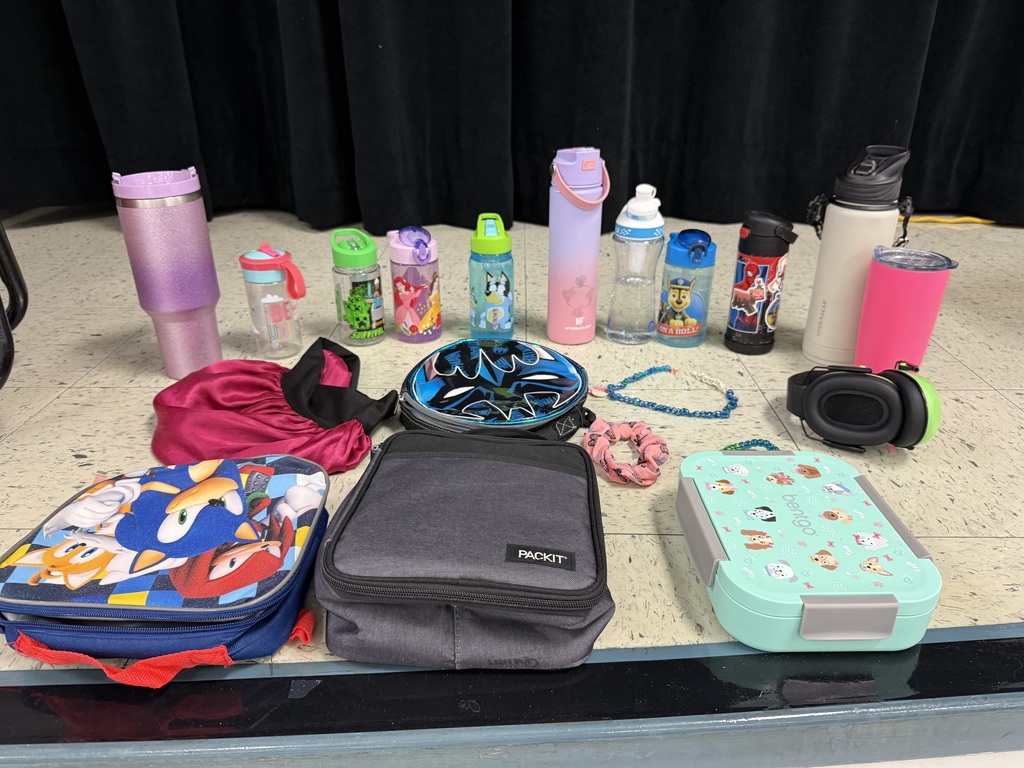 A collection of lost and found items from a school. Numerous children's jackets and hoodies are displayed on a clothing rack and on the floor. The collection also includes a variety of water bottles, lunch boxes, a hair bonnet, noise-canceling earmuffs, and jewelry laid out for identification.