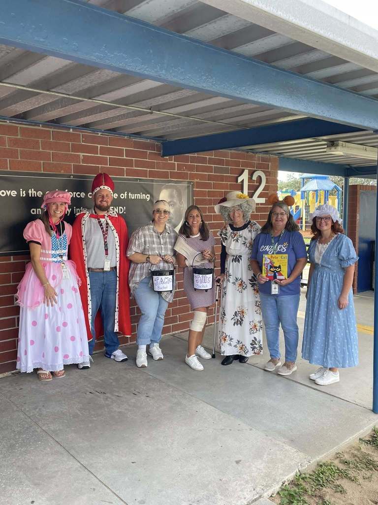 A group of adults stand outside a school building, dressed in playful costumes such as storybook characters and themed outfits. They are smiling and holding props, including small buckets and a book. The background shows a brick wall, a classroom door marked “12,” and part of a playground.